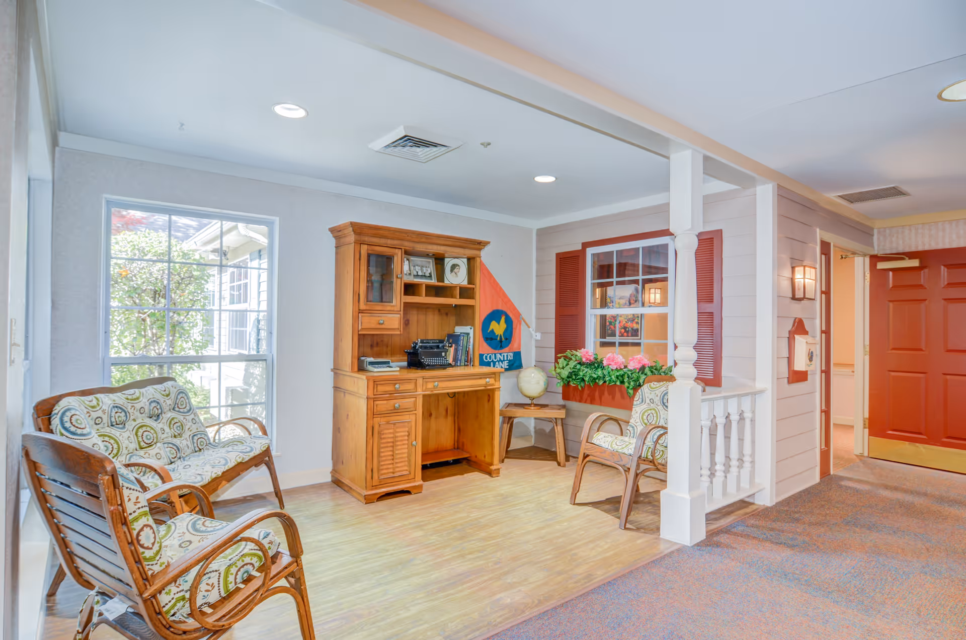 A cozy sitting area in a senior living facility with wooden chairs and a loveseat featuring patterned cushions. There is a wooden hutch with books, a typewriter, and decorative items, including a flag and a globe on a small table. A window with red shutters and a flower box with pink flowers adds a homey touch. The area has light-colored walls, wood flooring, and a red door in the background.