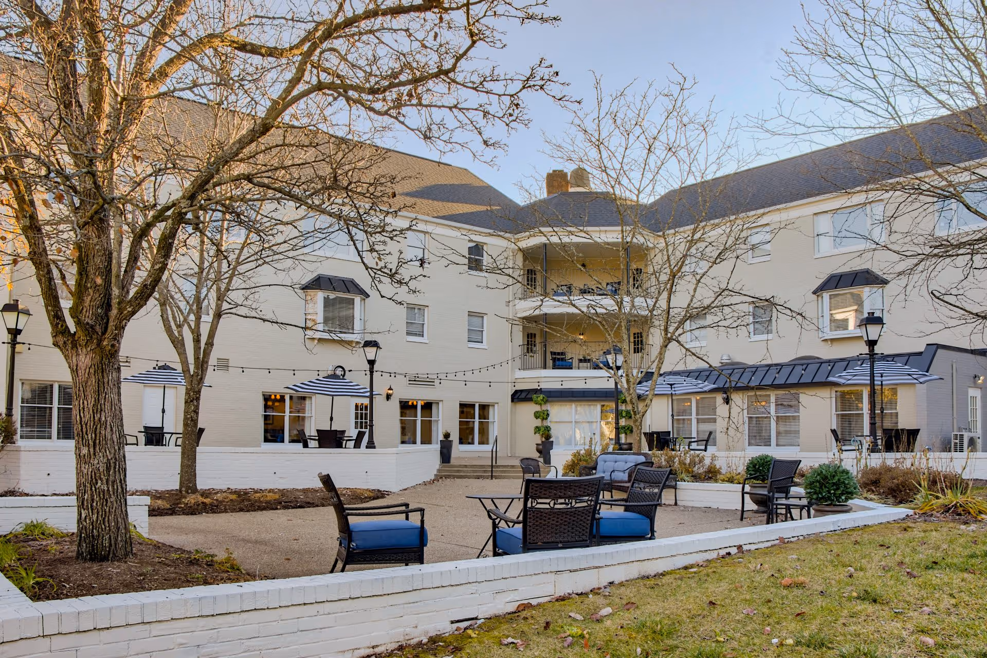 Outdoor courtyard of a multi-story residential building with seating areas, umbrellas, string lights, and leafless trees.