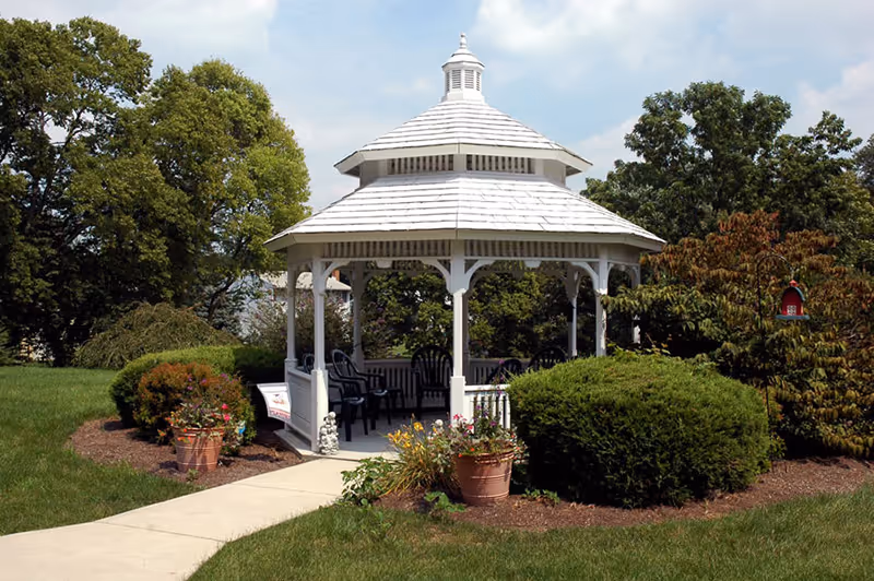 White wooden gazebo with a shingled roof surrounded by green bushes, potted flowers, and trees under a partly cloudy sky. There are several black chairs inside the gazebo and a concrete walkway leading up to it.