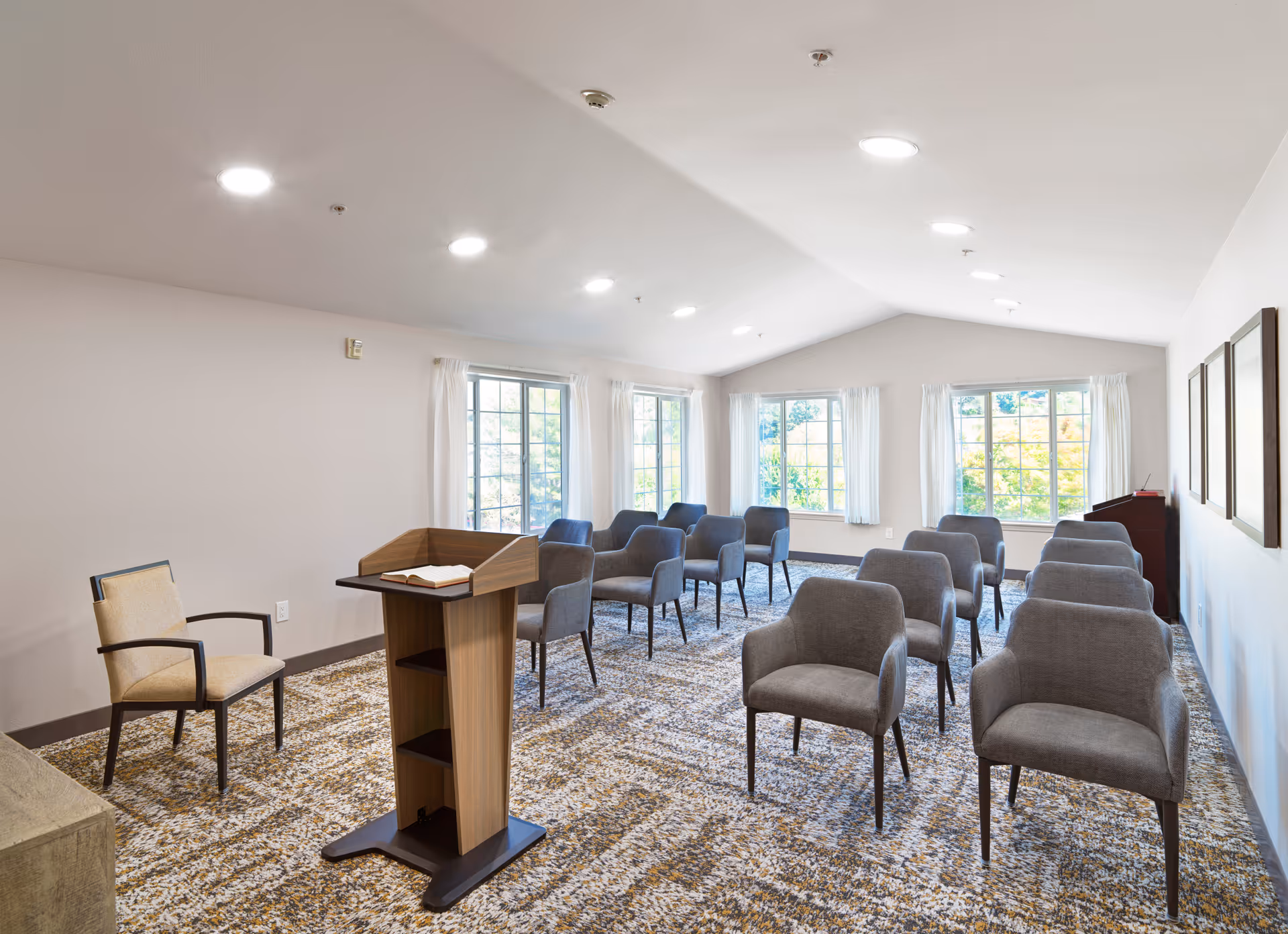 A bright room with multiple gray upholstered chairs arranged in rows facing a wooden podium with an open book on it. The room has large windows with white curtains letting in natural light, a patterned carpet, and three framed pictures on the right wall.