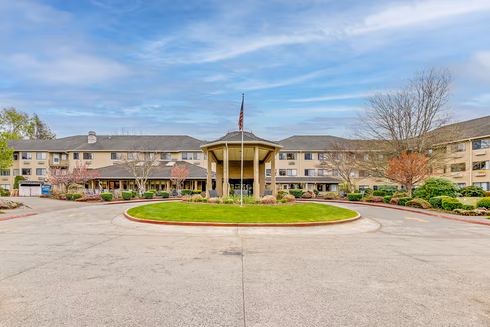 Front exterior view of Solstice Senior Living at Bellingham, showing a large three-story building with a covered entrance, a circular driveway with a grassy island in the center, and an American flag on a flagpole. The building is surrounded by landscaped bushes and trees under a partly cloudy sky.