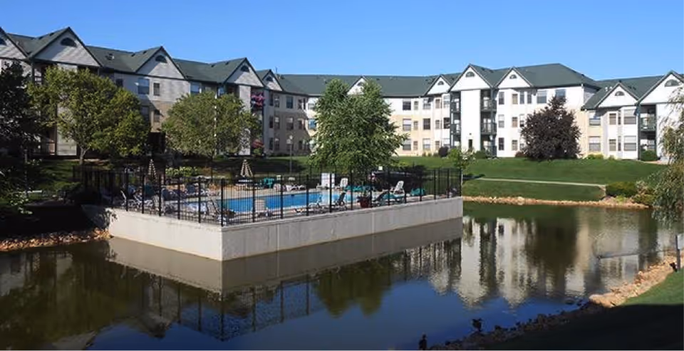 Exterior view of the Autumn Woods senior living complex showing a fenced pool on a platform over a pond with multi-story residential buildings behind it.
