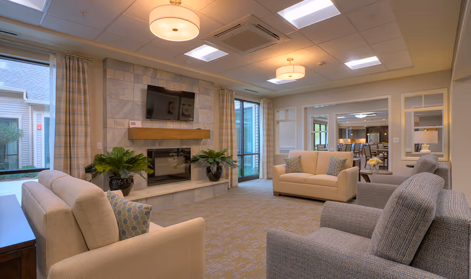 A cozy living room area in a senior living facility with beige and gray upholstered sofas and armchairs arranged around a stone fireplace with a mounted flat-screen TV above it. There are large windows with plaid curtains letting in natural light, potted plants on either side of the fireplace, and a view into an adjacent dining area with tables and chairs.