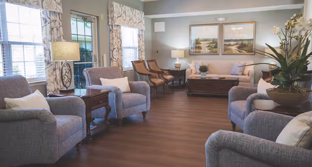 A cozy living room with several upholstered armchairs and wooden chairs arranged around wooden side tables and a coffee table. The room has large windows with patterned curtains, soft lighting from table lamps, and a large potted plant on a side table. Two landscape paintings hang on the wall above a sofa.