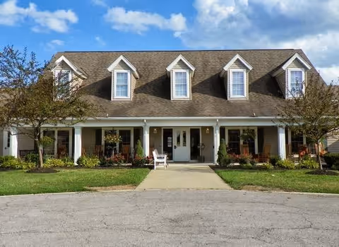 Front exterior view of a single-story residential building with a brown shingled roof featuring five dormer windows. The building has a covered porch with white columns, several rocking chairs, and is surrounded by green grass and small trees under a partly cloudy blue sky.