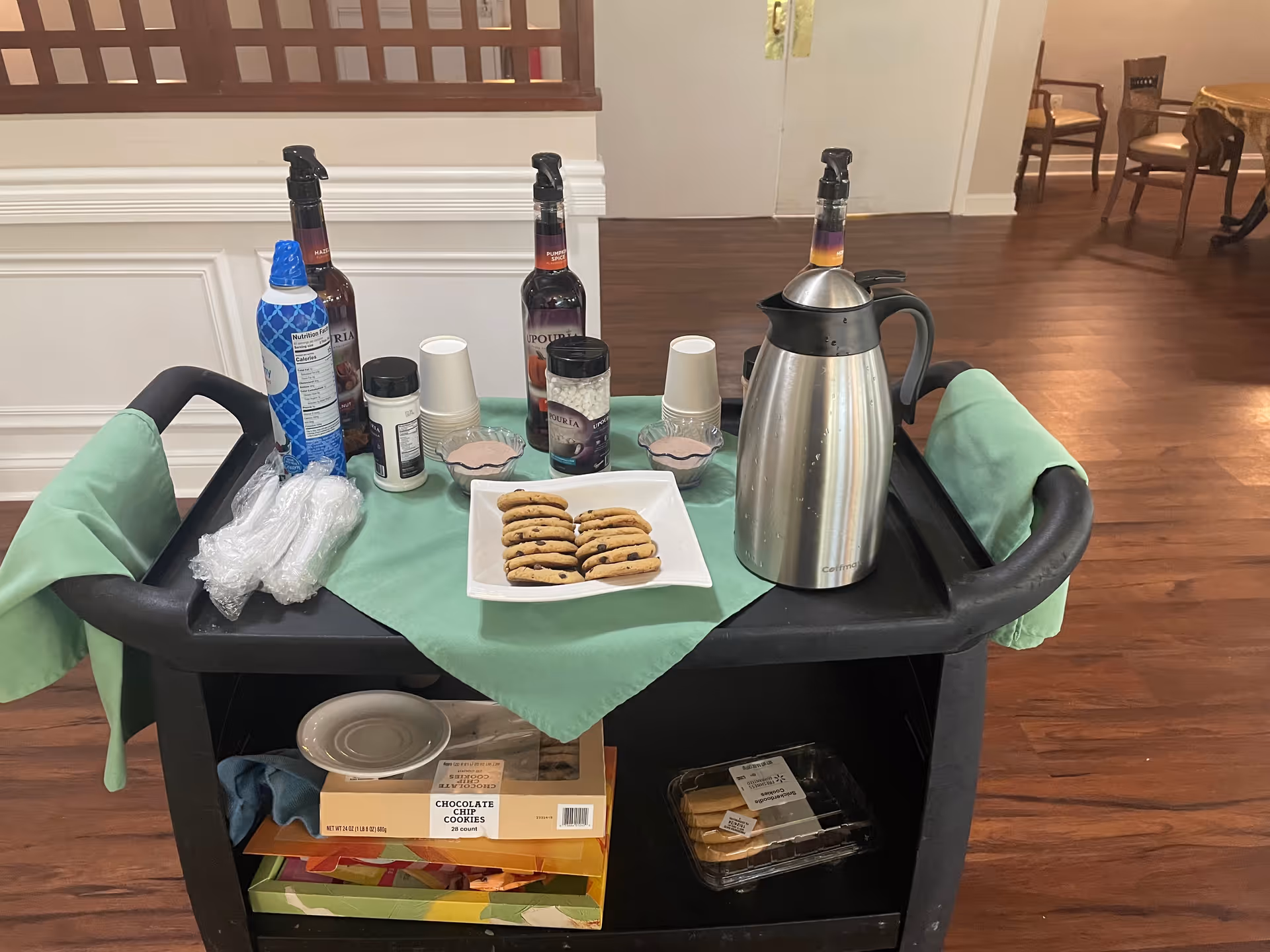 A black serving cart with green cloths draped over the handles, holding a plate of chocolate chip cookies, two bottles of syrup, a stainless steel coffee carafe, disposable cups, and various condiments. The cart is positioned on a wooden floor in a room with white walls and a wooden partition in the background.