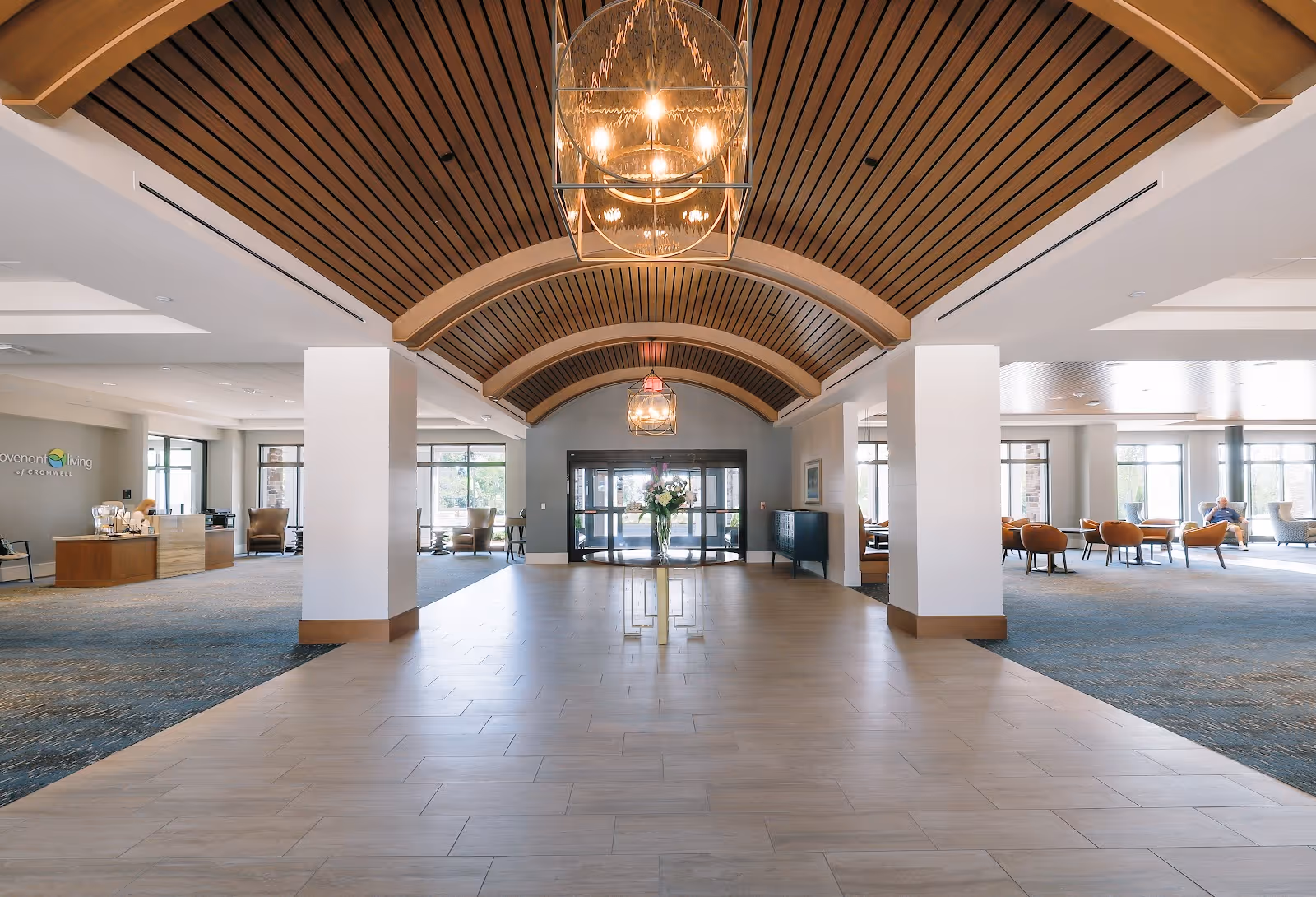 Spacious and well-lit interior lobby area with a wooden arched ceiling and modern hanging light fixtures. The floor is tiled in the center with carpeted seating areas on both sides, furnished with chairs and tables. Large windows allow natural light to fill the space, and a glass entrance door is visible at the far end. A flower arrangement is placed on a table in the middle of the tiled floor.