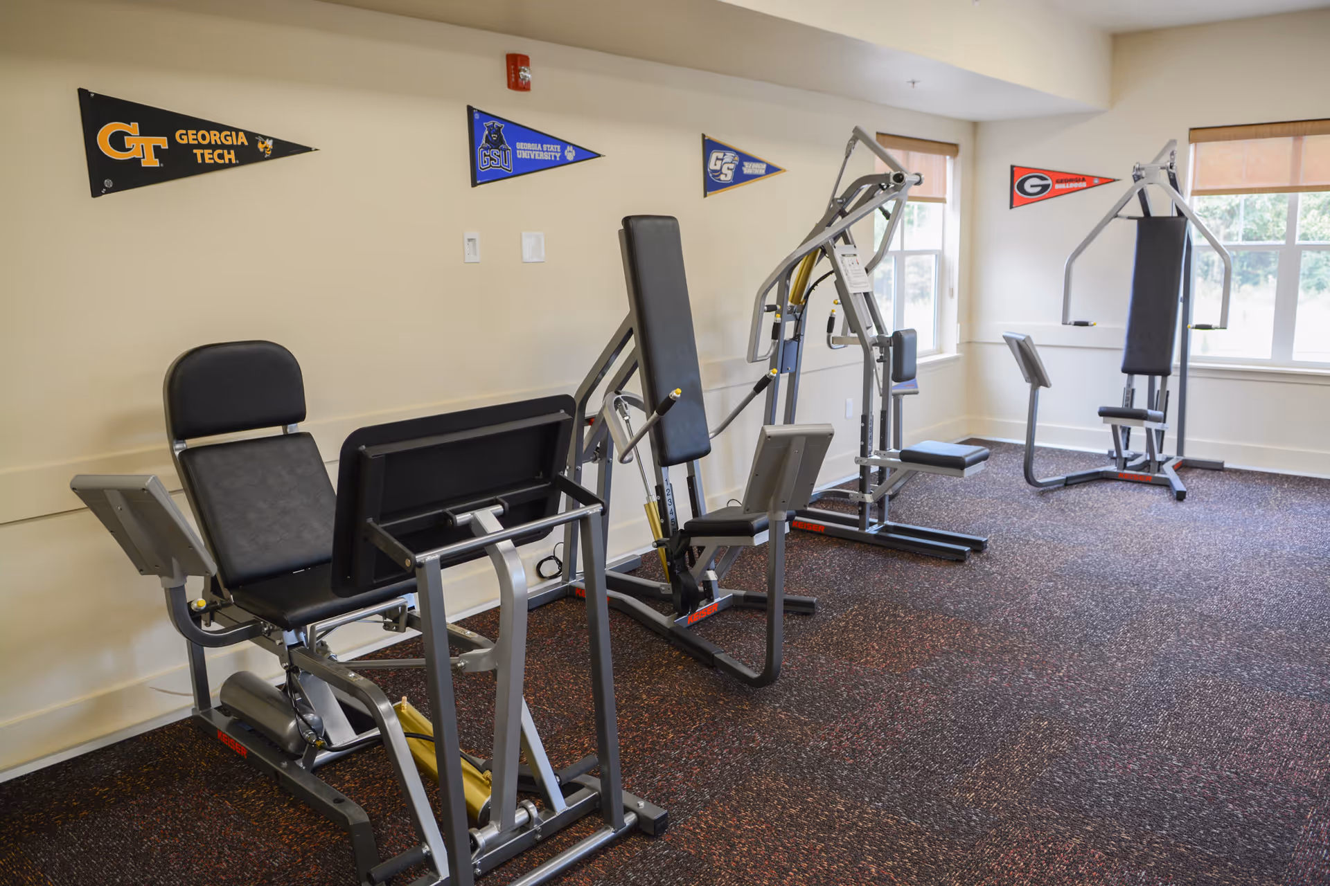 A small fitness room with several strength-training machines, windows, and college pennants on the wall.