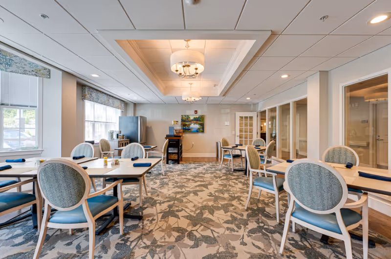 A bright and spacious dining room with several tables and chairs arranged neatly. The room features large windows with floral valances allowing natural light to fill the space. The ceiling has recessed lighting and two elegant chandeliers. There is a refrigerator and a small cabinet against the far wall, along with a colorful painting hanging above. The carpet has a leafy pattern, and the walls are painted in a light neutral color.