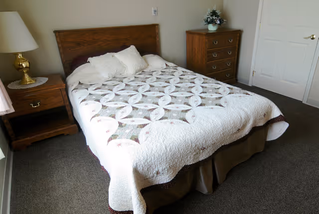 A bedroom with a double bed covered with a white quilt featuring a circular pattern. The bed has two pillows and a wooden headboard. To the left of the bed is a wooden nightstand with a brass lamp and a doily. To the right is a wooden chest of drawers with a floral arrangement on top. The room has beige walls, a carpeted floor, and a closed white door.