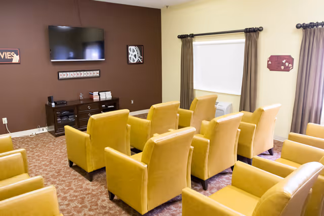 A small movie viewing room with two rows of yellow armchairs facing a wall-mounted flat screen TV. The room has a brown accent wall with movie-themed decorations and a wooden media console below the TV. There are windows with brown curtains on the adjacent wall.