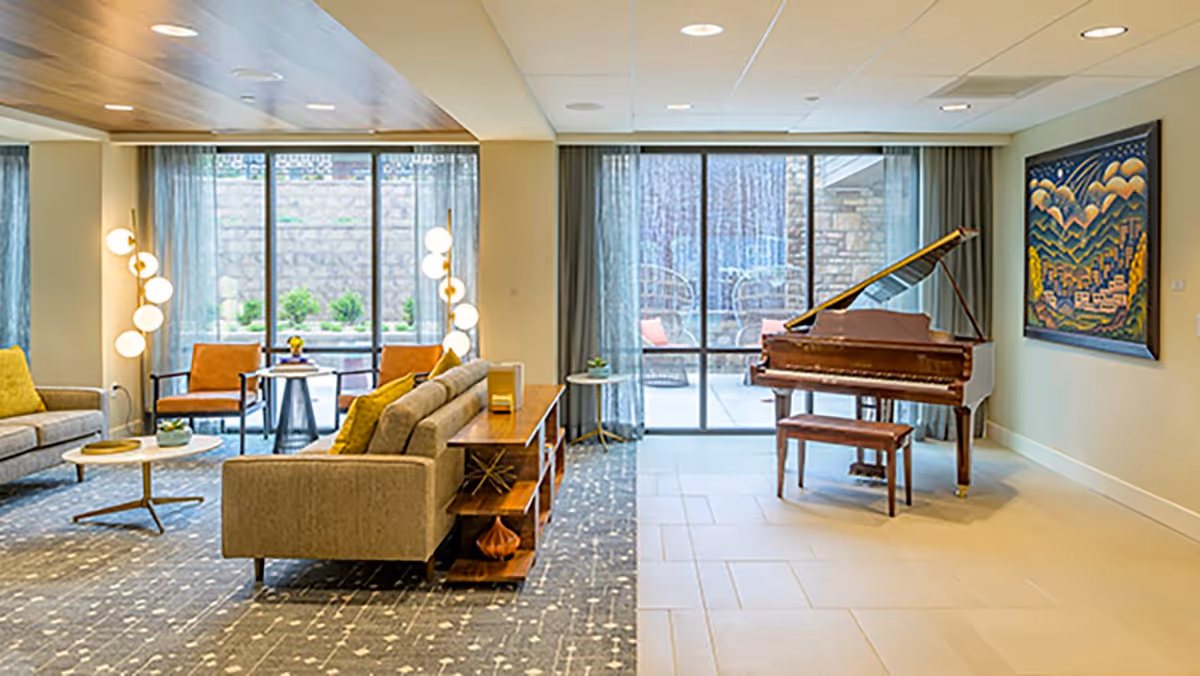 A spacious senior living facility lounge area featuring comfortable seating with sofas and armchairs, a grand piano with a matching bench, modern floor lamps, large windows with sheer curtains allowing natural light, and a colorful framed artwork on the wall.