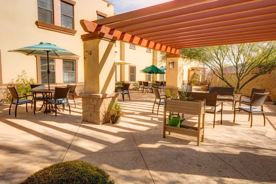Outdoor patio area at Tucson Place at Ventana Canyon with several tables and chairs under a wooden pergola. There are umbrellas over some tables, potted plants, and desert landscaping with trees and bushes surrounding the space.