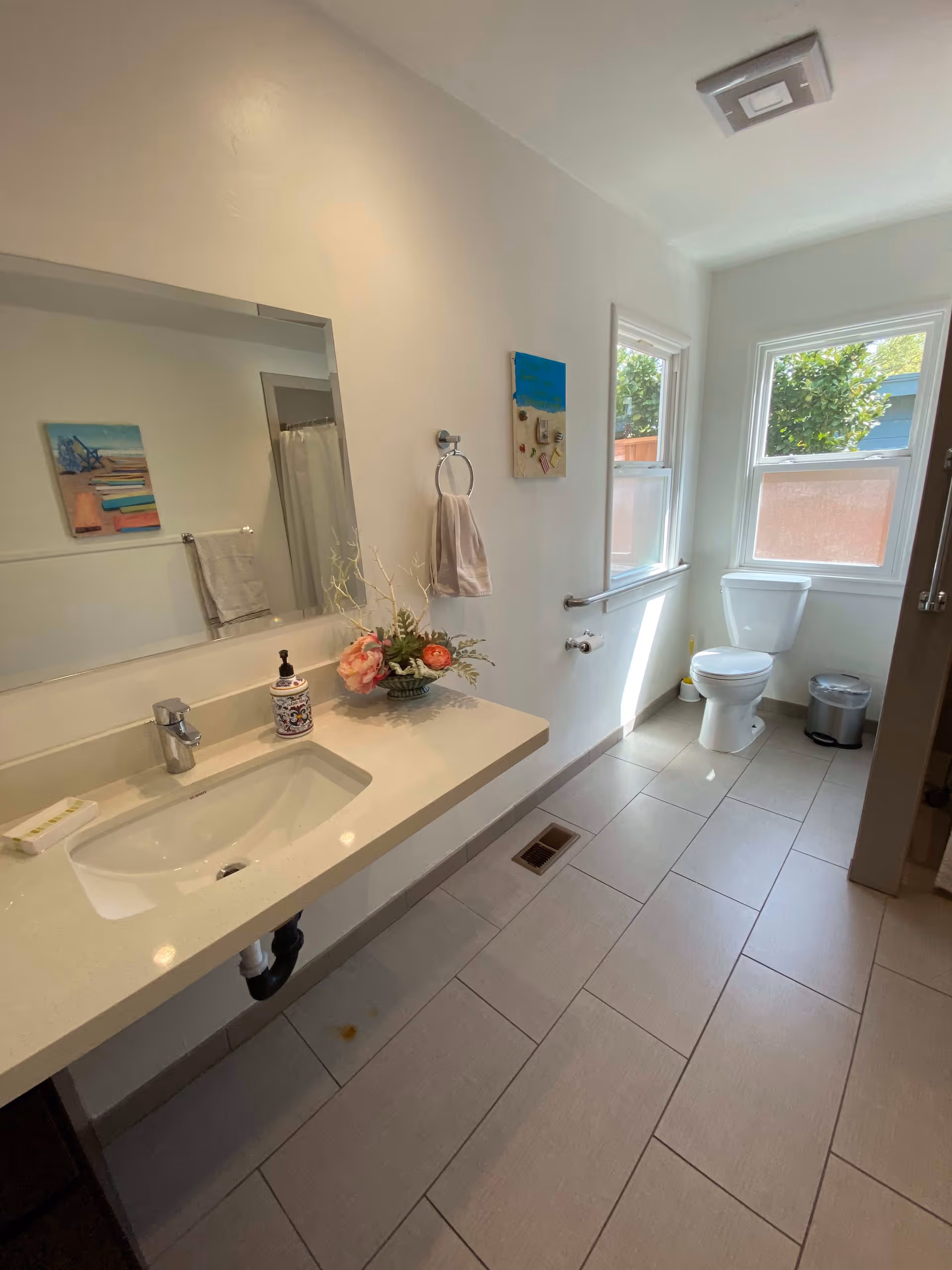 A clean and bright bathroom with a large mirror above a white countertop sink. There is a decorative flower arrangement and a soap dispenser on the counter. A towel hangs on a ring on the wall next to the sink. The bathroom has two windows letting in natural light, a toilet, a small trash can, and a shower with a curtain partially visible. The floor is tiled in light gray.