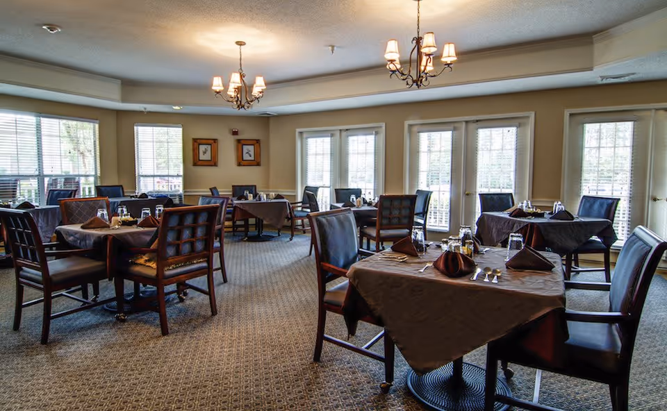 Sunlit dining room with multiple tables set with tablecloths, chairs, and chandeliers.