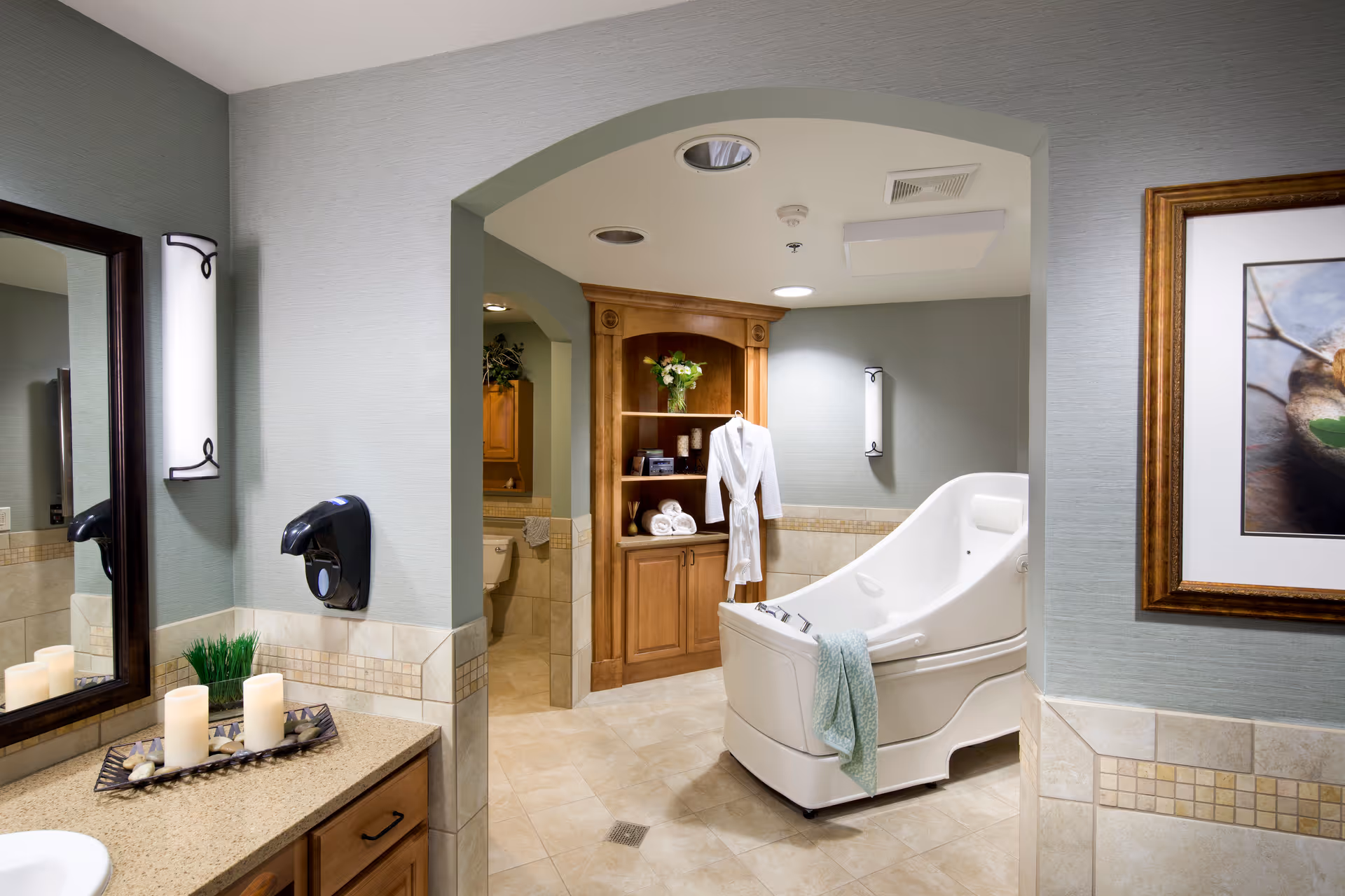 A spacious, well-lit bathroom featuring a large white therapeutic bathtub with a light blue towel draped over the side. The room has beige tiled floors and walls with decorative tile accents. There is a wooden cabinet with shelves holding a vase of flowers, rolled towels, and candles. A white bathrobe hangs on the cabinet. A countertop with candles and a small plant is visible, along with a wall-mounted soap dispenser and a large mirror. The walls are painted a soft greenish-gray color, and there is framed artwork on the wall.