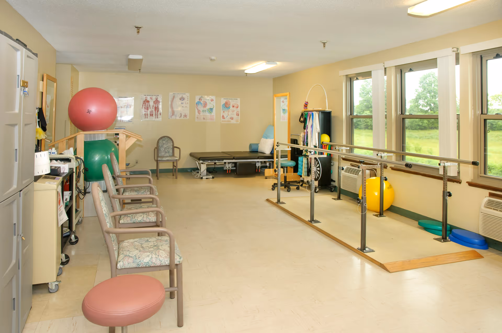 A physical therapy room with parallel bars for walking exercises, exercise balls, chairs lined up against the wall, anatomical charts on the wall, and large windows letting in natural light.
