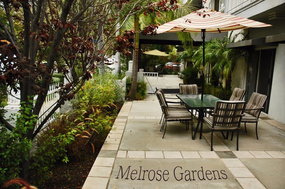 Outdoor patio area with a glass-top table and six striped cushioned chairs under a large striped umbrella. The patio is surrounded by lush greenery and trees, with a building on the right side. The words 'Melrose Gardens' are visible on the tiled ground near the table.