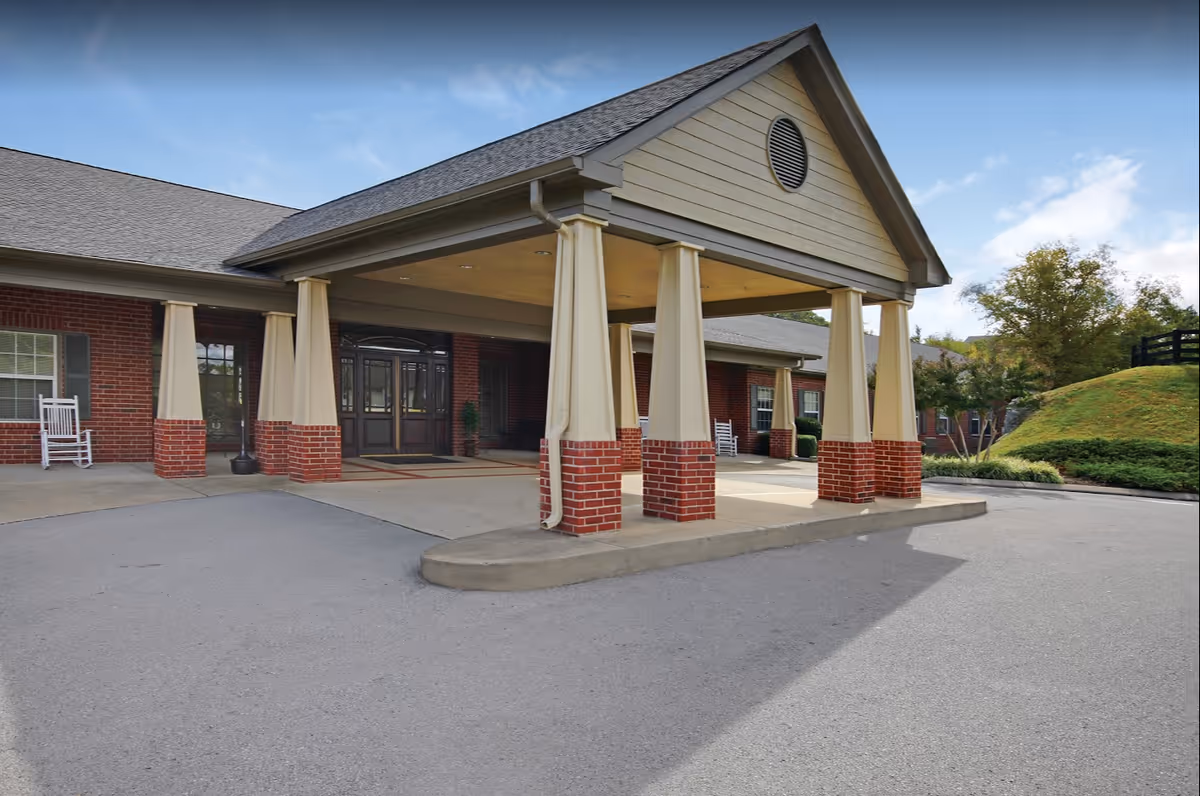 Entrance of a senior living facility with a covered drop-off area supported by columns with brick bases. The building has a brick exterior with windows and white rocking chairs on the porch. There is a driveway and some greenery in the background under a partly cloudy sky.