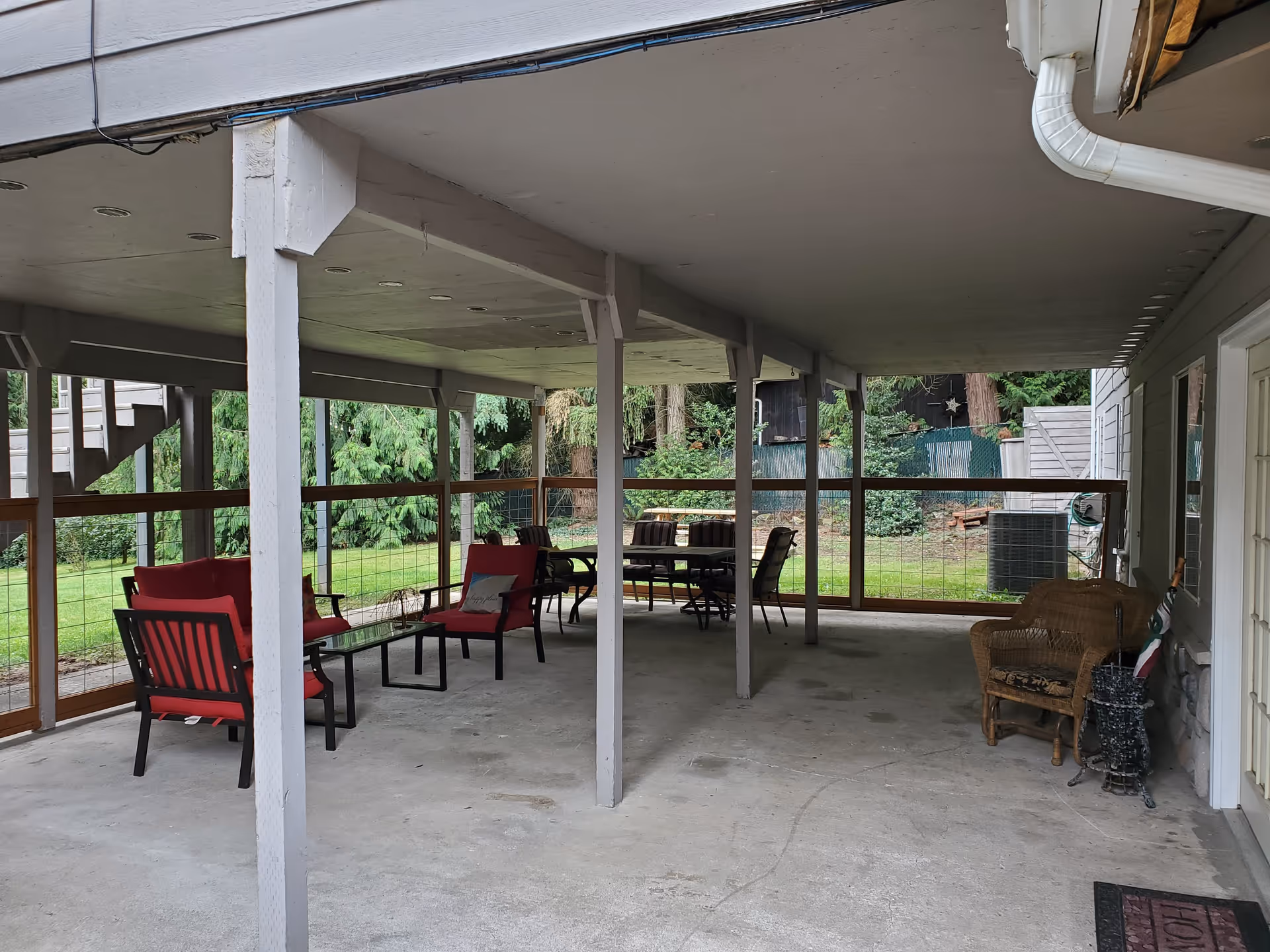 Covered outdoor patio area with several seating arrangements including red cushioned chairs around a glass table and a wicker chair near the wall. The patio is supported by white wooden beams and overlooks a green grassy yard with trees and a fence.
