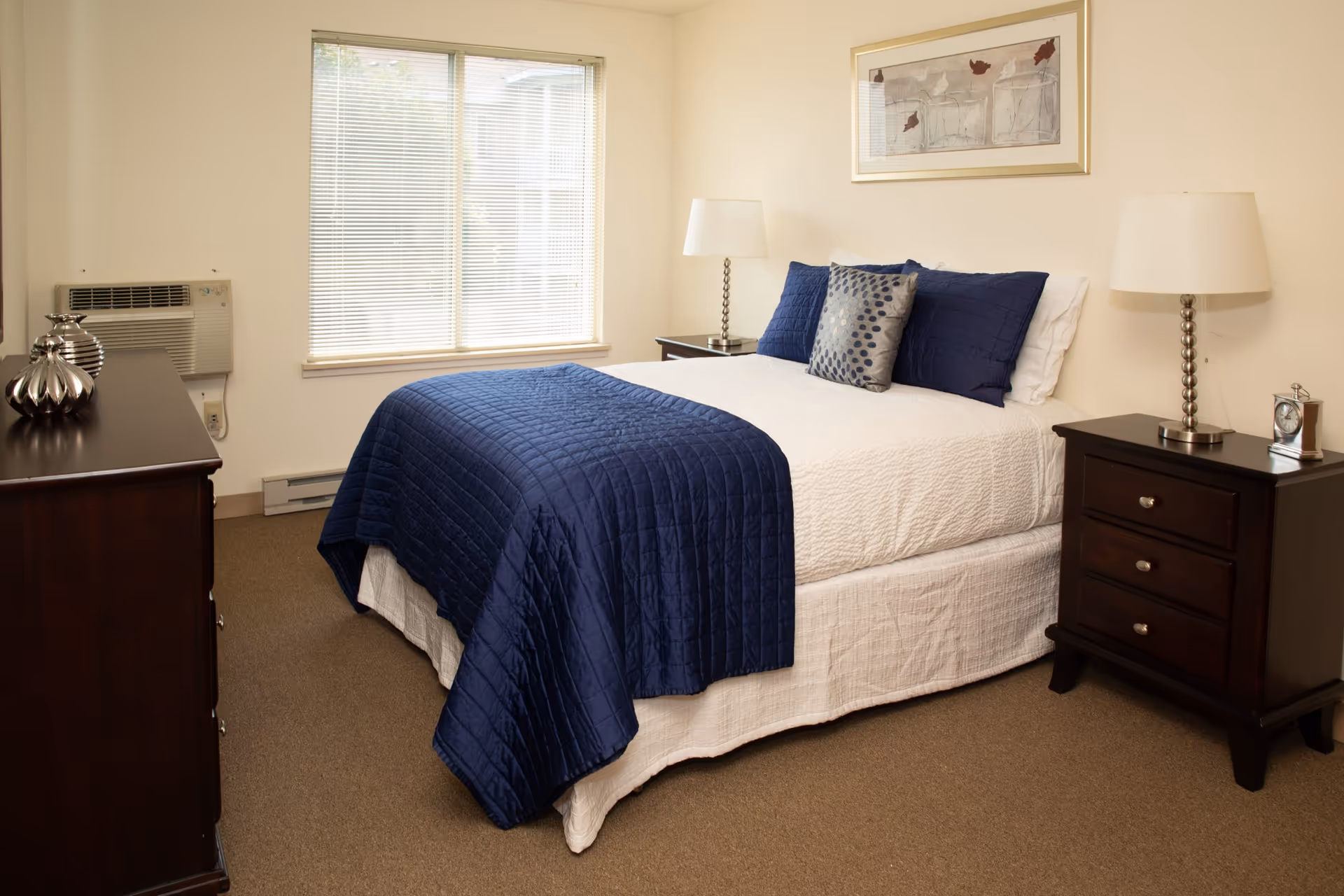 A neatly made bed with white sheets and a navy blue quilt and pillows in a senior living bedroom. The room features beige walls, a window with blinds, a dark wooden dresser with decorative items, and a matching nightstand with a lamp and a clock. A framed artwork hangs above the bed.