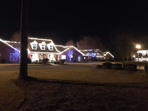 Nighttime view of a residential-style building decorated with white string lights along the roofline and blue lights on trees in front. The building is surrounded by a lawn and some bushes, with a streetlamp illuminating part of the scene.