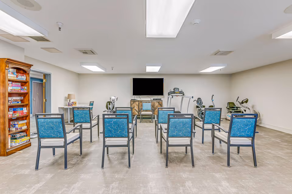 A spacious activity room with a row of blue cushioned chairs facing a large flat-screen TV mounted on the wall. To the left, there is a wooden shelf filled with board games and a small table with a lamp. On the right side of the room, there are several pieces of exercise equipment including a treadmill and stationary bikes. The room has a light-colored carpet and white walls with ceiling lights.
