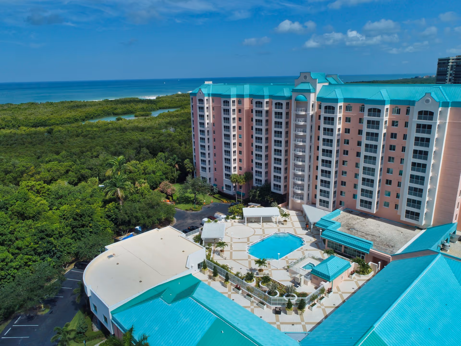 Aerial view of Premier Place senior living facility featuring a large multi-story building with turquoise roofs, a central swimming pool, and surrounding greenery with a distant view of the ocean under a blue sky with scattered clouds.