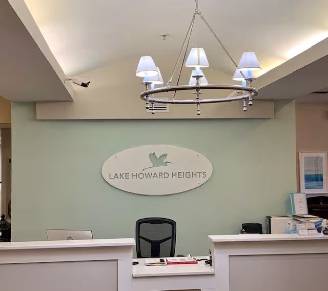 Reception desk area with a black office chair behind it, a chandelier with five lamps hanging from the ceiling, and a wall sign that reads 'Lake Howard Heights' with a bird logo above the text. The wall is painted light green and there are office supplies and papers on the desk.