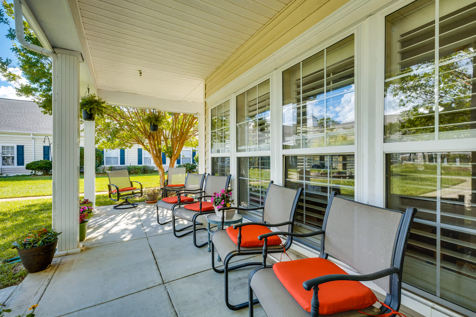 Covered outdoor patio area with several chairs featuring red cushions arranged along the side of a building with large windows. Potted plants and hanging planters decorate the space, and a green lawn with trees and other buildings is visible in the background under a blue sky with some clouds.