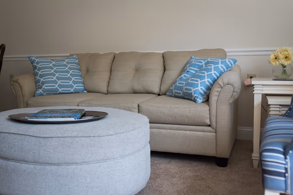 Beige sofa with blue patterned throw pillows, a round gray ottoman with books on a tray, and a small side table with flowers in a living room.