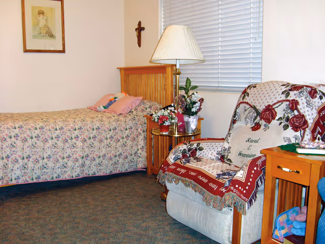 A cozy bedroom with a single bed covered in a floral bedspread, a wooden headboard, and a pink pillow. Next to the bed is a wooden nightstand with a brass lamp and several flower arrangements. A comfortable armchair draped with a decorative throw blanket and a personalized pillow sits nearby. The room has a carpeted floor, a window with closed blinds, a framed picture on the wall, and a small wooden side table with a drawer and some items on it.