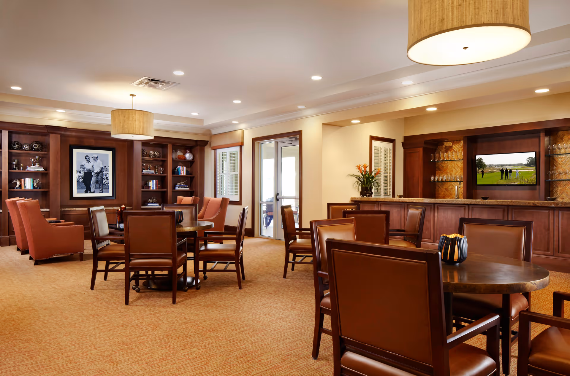 A spacious, well-lit common area with several round tables surrounded by brown leather chairs. The room features built-in wooden shelves with decorative items and a large black and white photo on the wall. There is a bar area with glassware and a TV mounted above it. The carpet is beige, and the ceiling has recessed lighting and a large hanging lamp.