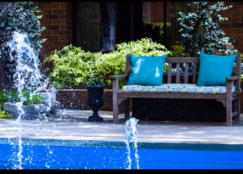 Outdoor seating area with a wooden bench featuring two turquoise cushions and a patterned seat cushion, situated near a water fountain with water jets spraying upwards. Green shrubs and plants are visible in the background along with a brick wall.