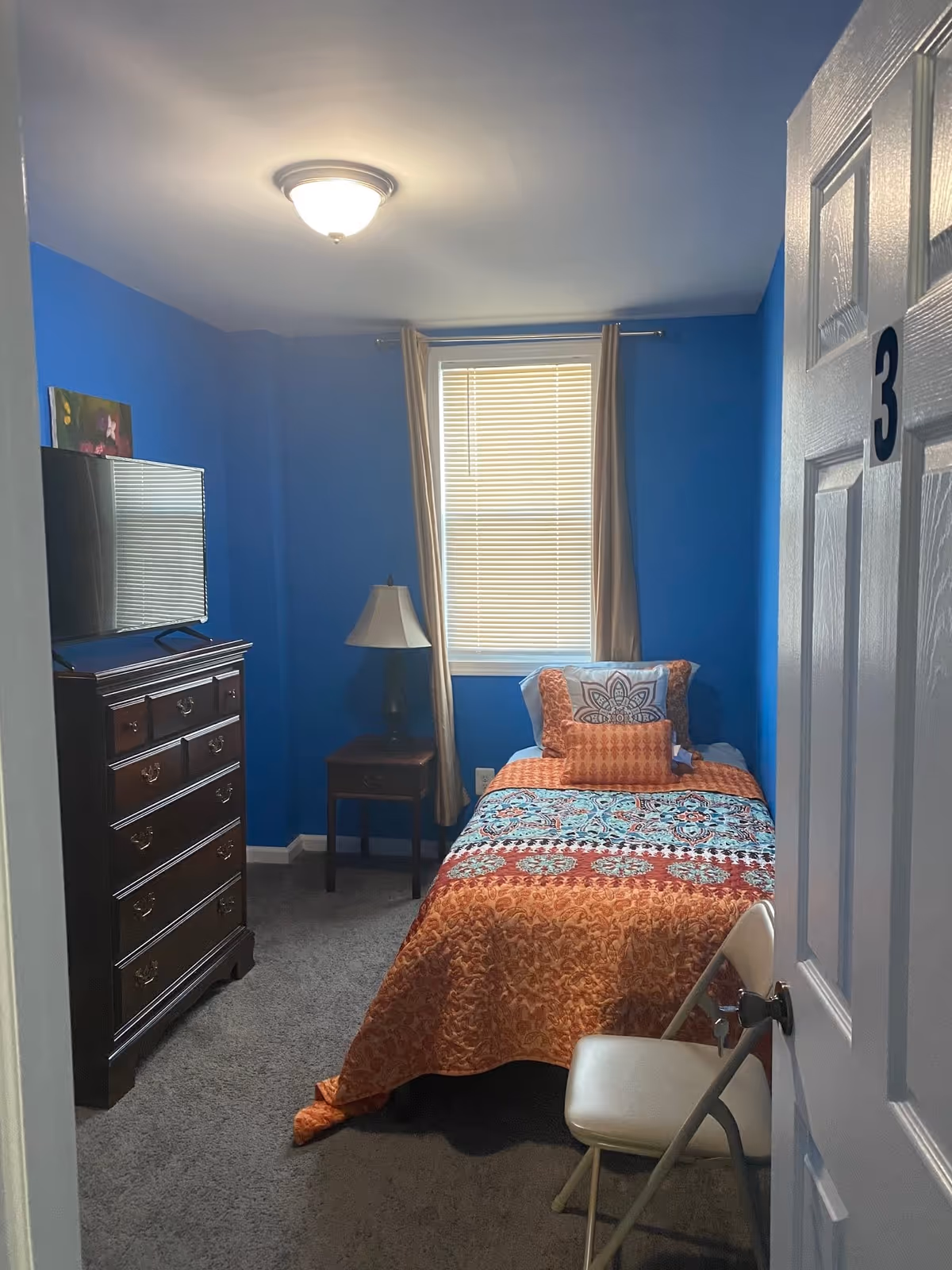 A small bedroom with bright blue walls and a single bed covered with an orange and blue patterned quilt. There is a wooden dresser with a flat-screen TV on top, a small wooden nightstand with a lamp, and a white folding chair near the door. The window has beige curtains and closed blinds. The door has the number 3 on it.