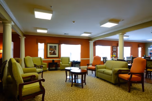 Well-lit senior living facility common lounge with upholstered chairs, sofas, columns, and coffee tables.