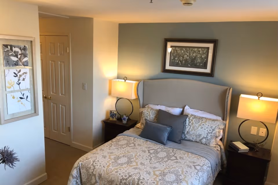 A cozy bedroom with a neatly made bed featuring patterned bedding and multiple pillows. The bed has a beige upholstered headboard. On either side of the bed are dark wooden nightstands with modern lamps emitting warm light. A framed floral artwork hangs above the bed, and another piece of framed botanical art is on the adjacent wall near a closed door.