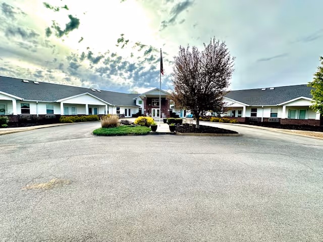Front entrance of a single-story assisted living facility with a circular driveway, central flagpole, and landscaped island.