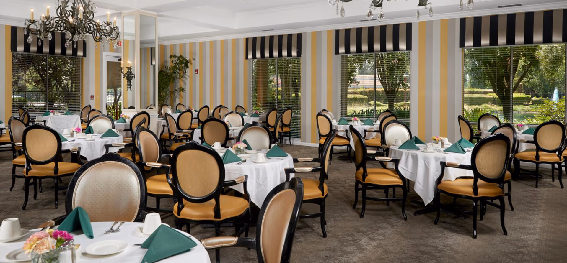 Bright dining room with round tables set with white linens and green napkins, upholstered chairs, chandeliers, and large windows overlooking a garden.
