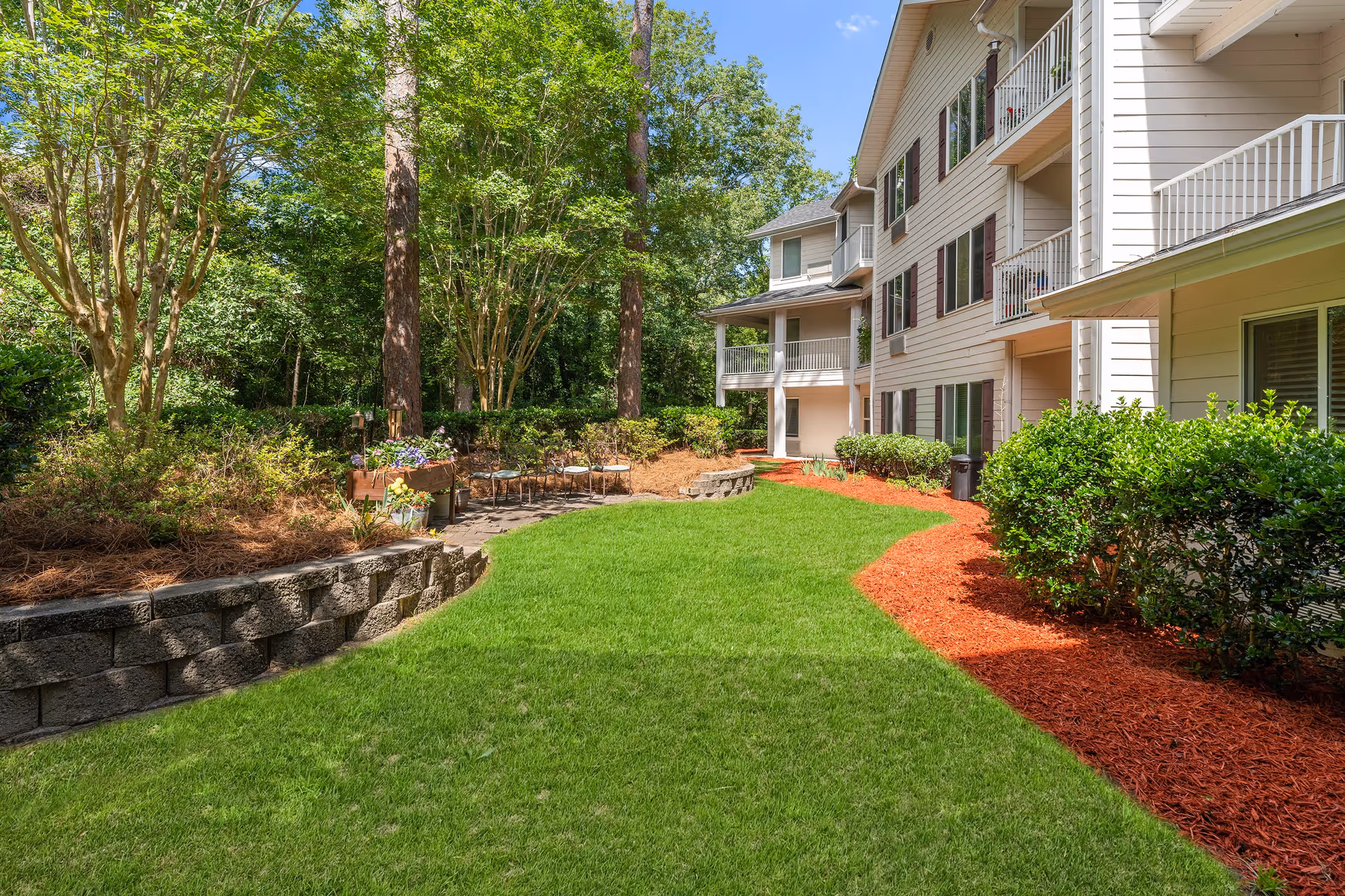 A well-maintained outdoor garden area beside a multi-story residential building with balconies. The garden features a lush green lawn, a stone retaining wall, several trees, shrubs, and a small seating area with chairs and a planter box filled with flowers. The building has beige siding and white railings on the balconies.