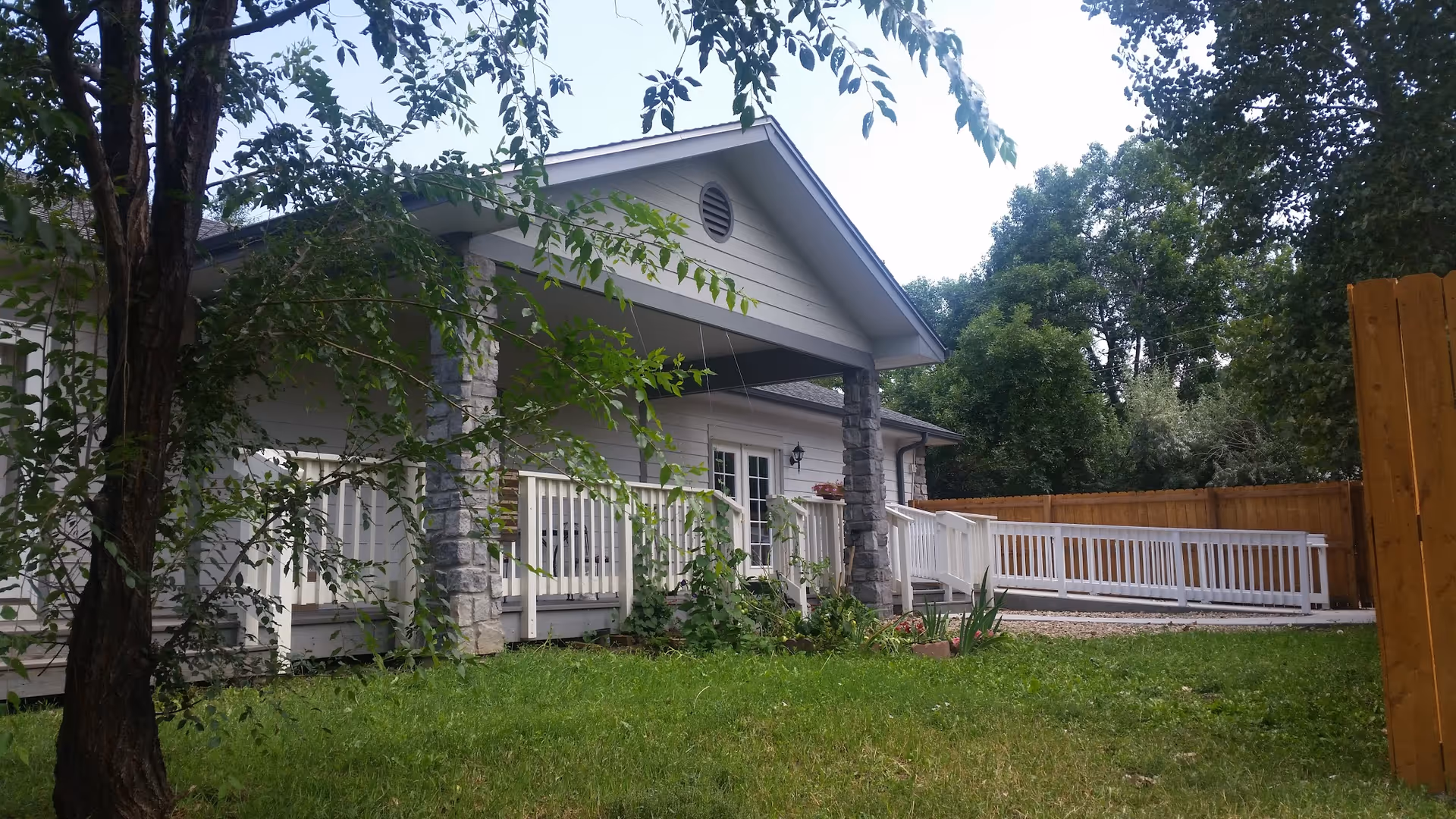 Exterior view of a single-story building with a covered porch supported by stone pillars. The porch has white railings and a wheelchair accessible ramp. The building is surrounded by green grass, trees, and a wooden fence in the background.