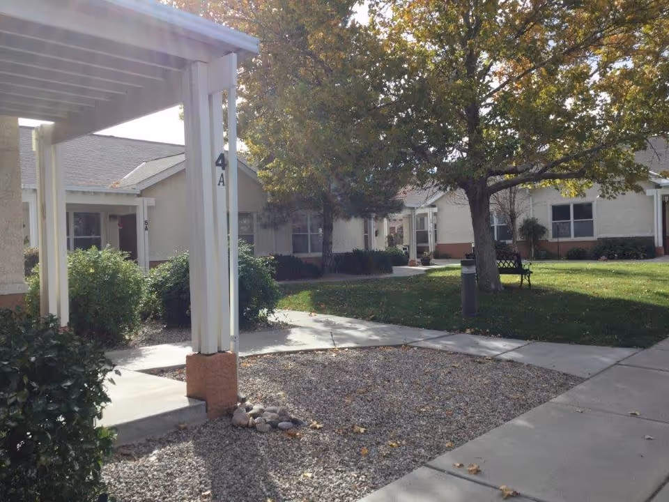 Outdoor courtyard area of a senior living facility with a large tree, green grass, bushes, a bench, and multiple single-story buildings with beige walls and brown trim. There is a covered walkway with a post labeled '4A' and paved walkways connecting the buildings.