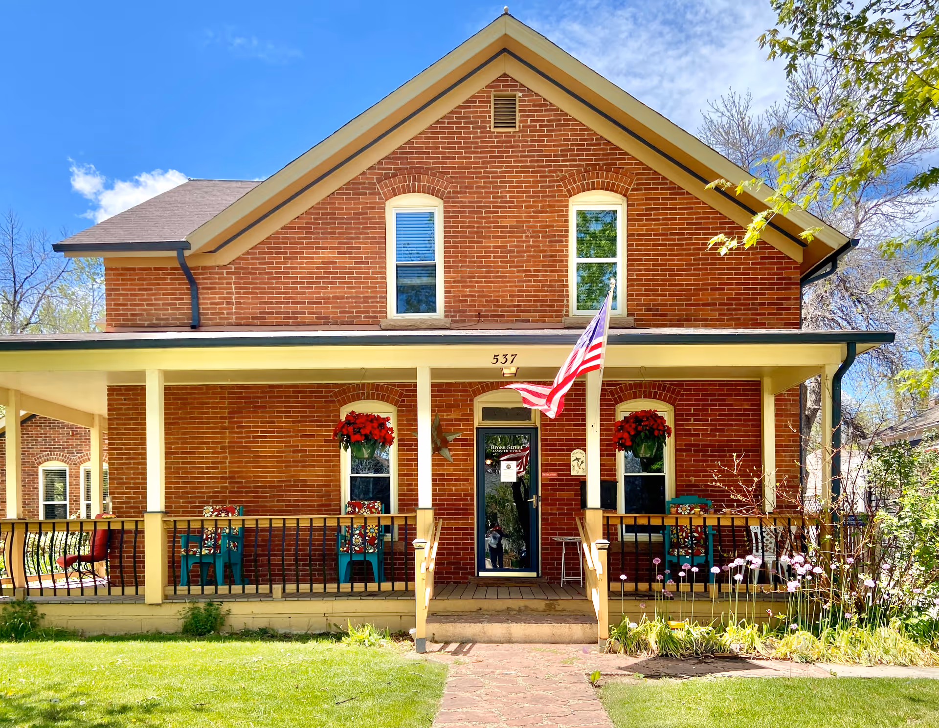 Front exterior view of a two-story brick building with a covered porch. The porch has several chairs with floral cushions, hanging flower baskets, and an American flag mounted near the entrance door. The building number 537 is displayed above the door, and there is a well-maintained lawn and garden in front.