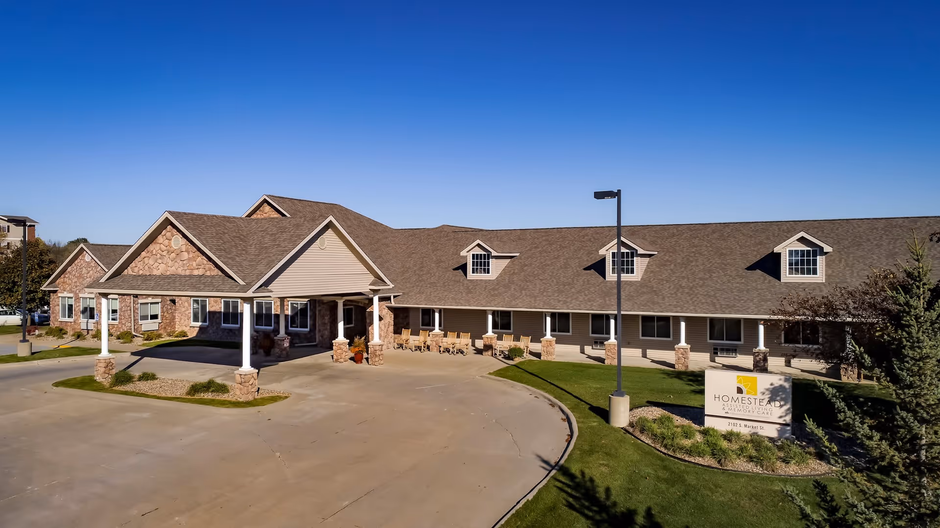 Exterior front view of Homestead Assisted Living & Memory Care of Oskaloosa, showing a single-story building with a covered entrance, stone and siding facade, multiple windows, a driveway, and a sign with the facility name on a landscaped lawn under a clear blue sky.