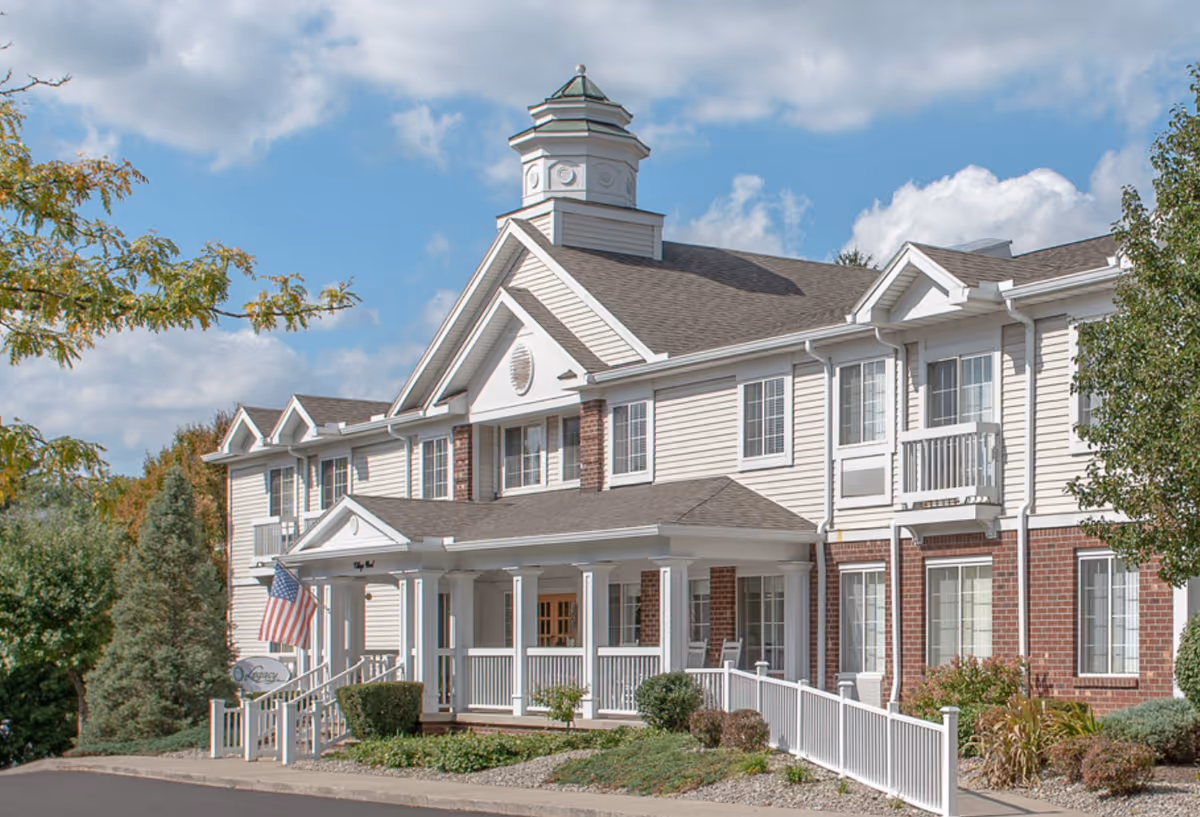 Exterior view of a two-story senior living facility building with white siding and red brick accents. The building features a covered entrance with white columns, an American flag, and a small sign that reads 'Legacy'. There are bushes and trees surrounding the building under a partly cloudy blue sky.