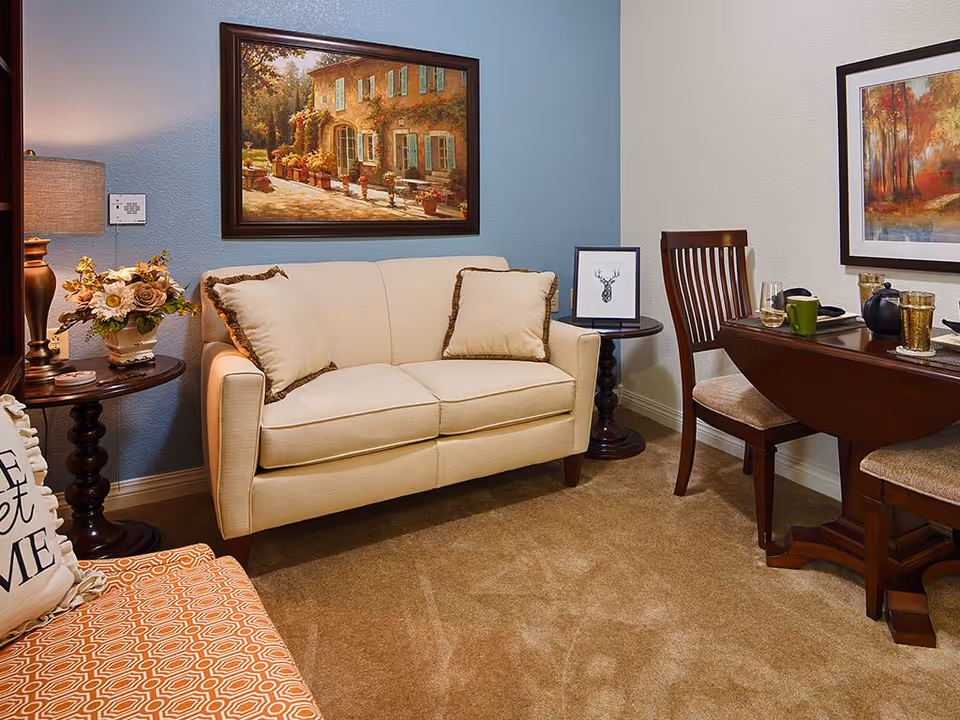 Cozy living room area with a beige loveseat adorned with two matching pillows, a wooden side table with a floral arrangement and lamp, a wooden dining table set with chairs, cups, and a teapot, and framed artwork on the walls.