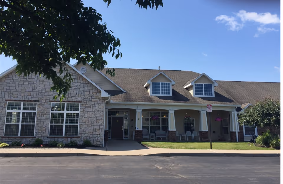 Front exterior view of a single-story senior living community building with stone and siding facade, large windows, a covered porch with chairs, hanging flower baskets, and a clear blue sky above.