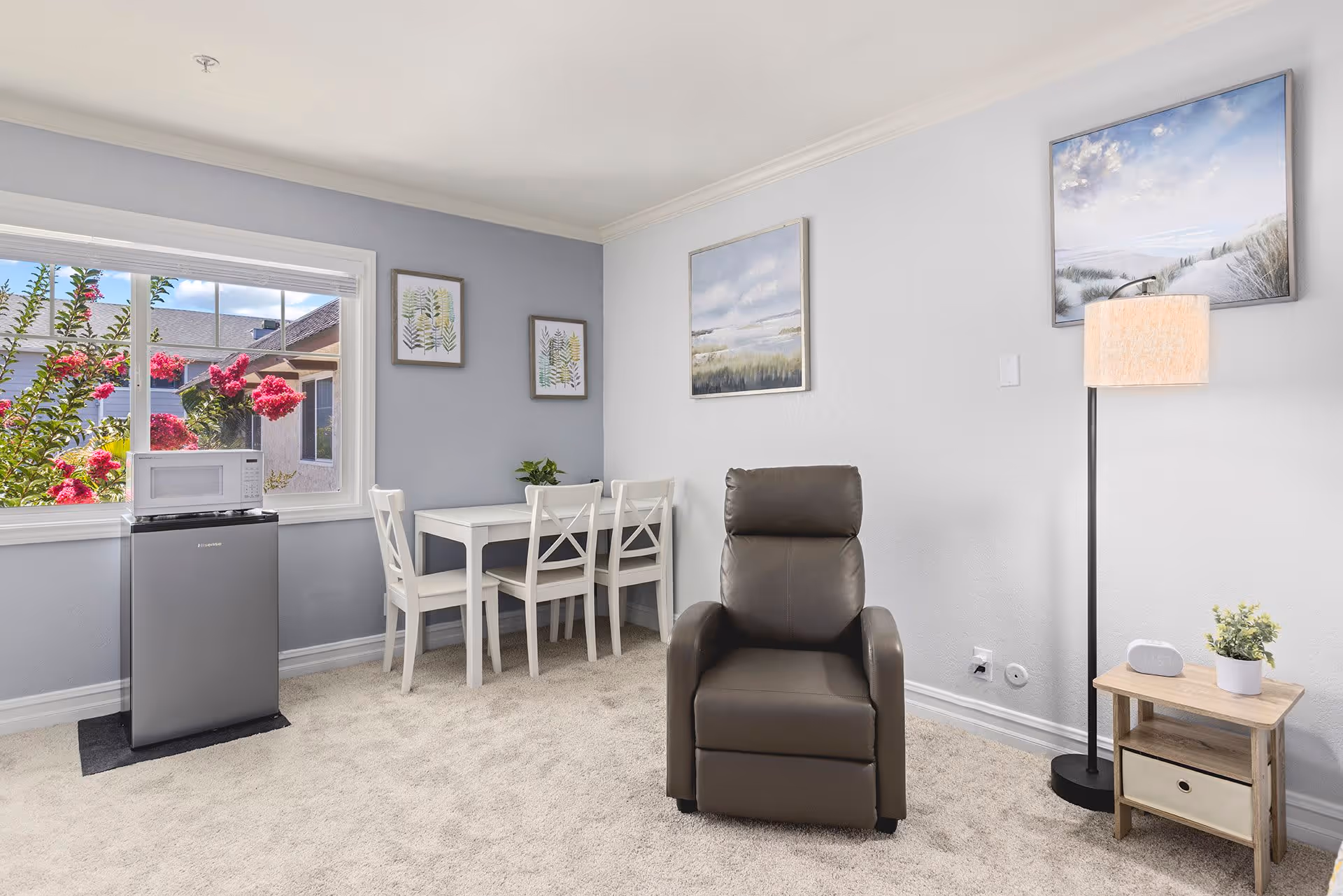 A bright and cozy living area with a gray recliner chair, a small wooden side table with a plant and a clock, a tall floor lamp, and a white dining table with three chairs. There is a window showing pink flowers outside, and three framed pictures hanging on the light gray walls.