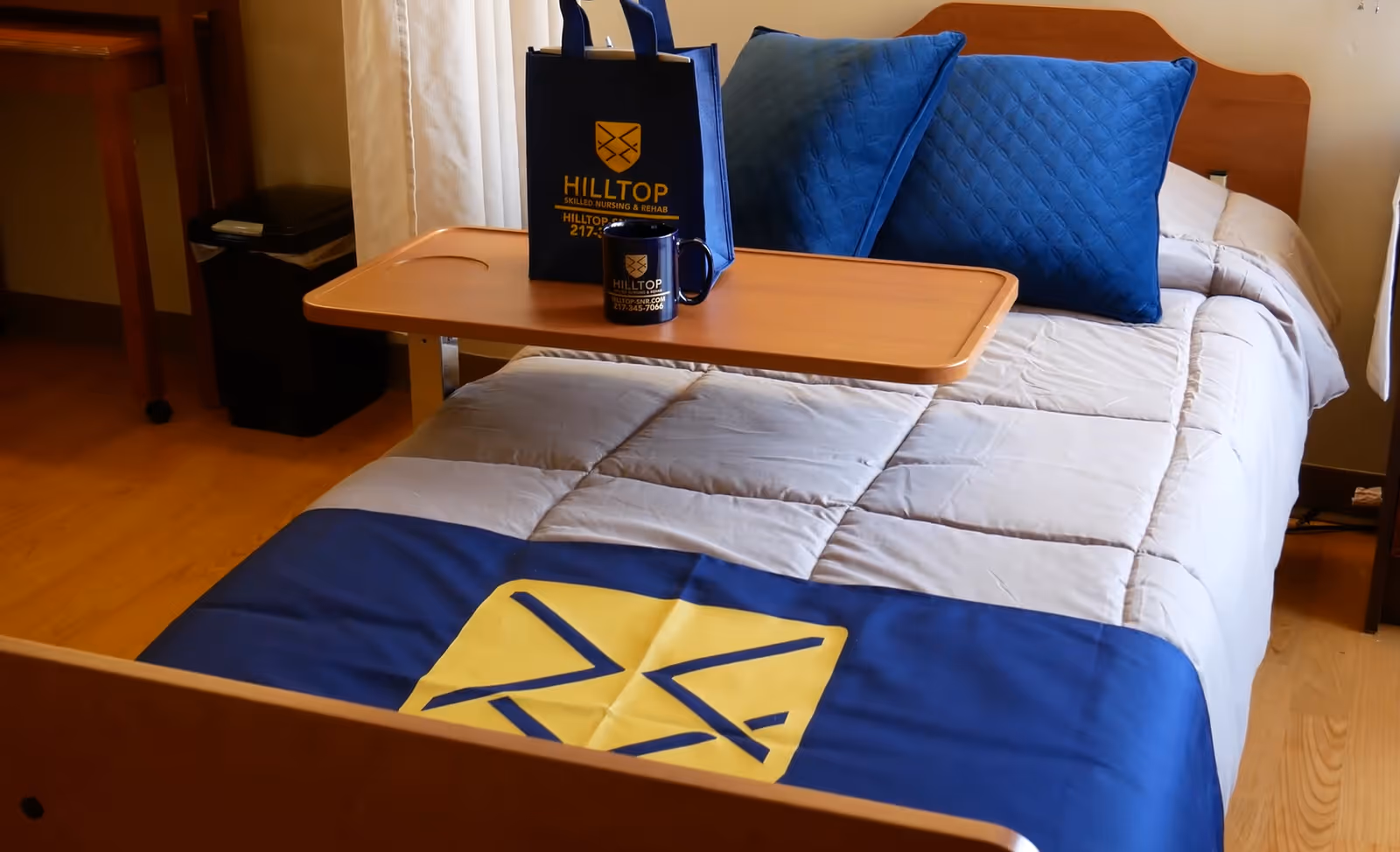 A neatly made single bed in a skilled nursing facility room with a gray and blue comforter, two blue pillows, and a wooden overbed table holding a blue tote bag and matching mug with the Hilltop Skilled Nursing & Rehab logo.