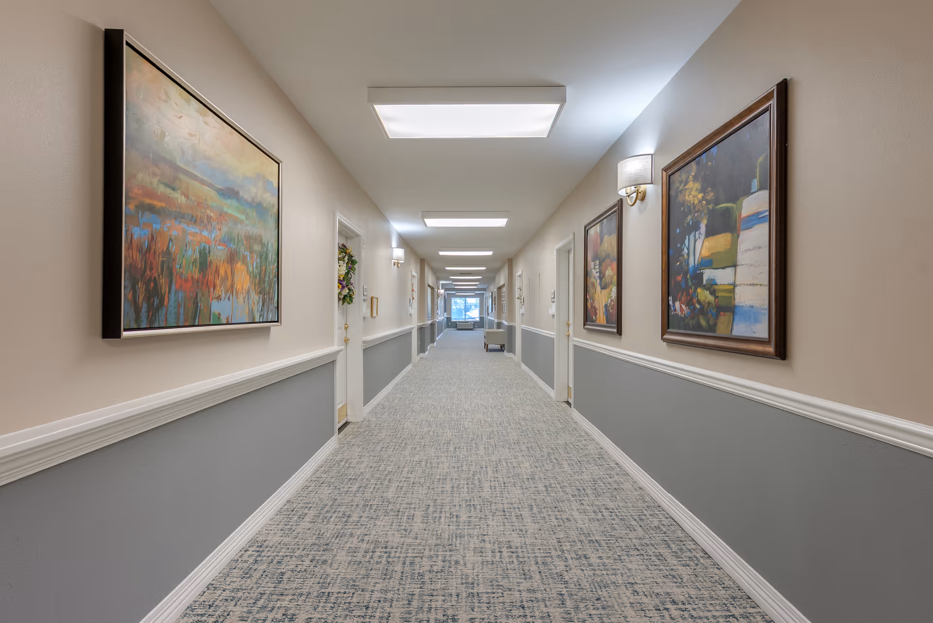 Well-lit carpeted hallway lined with framed artwork and doors in a senior living facility.